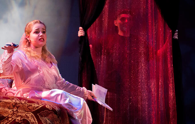 An actor sits to the left, drapped in fine shiny silks, and speaks to the audience. To the right of her is a glistening, red-tinted, warm curtain, with a prop holder behind it.