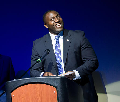 A speaker looks up at an off-screen teleprompter, smiling widely.