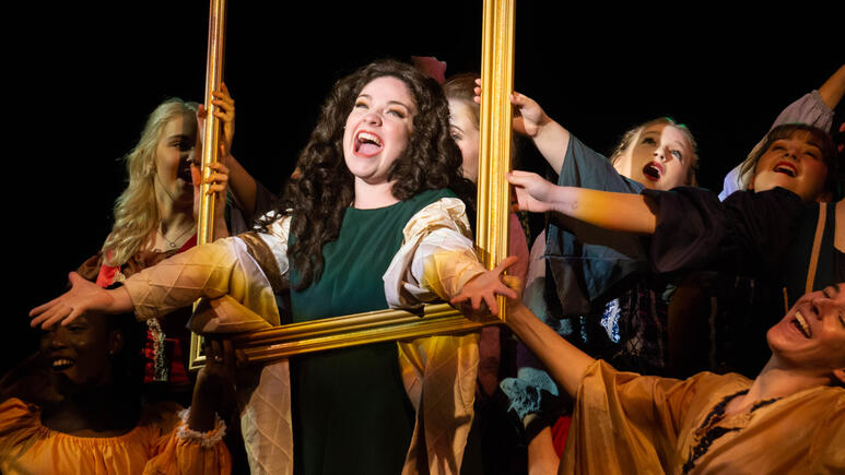 Actors stand, arms spread, reaching through an empty photo frame toward the audience. Their eyes are alight while they sing.