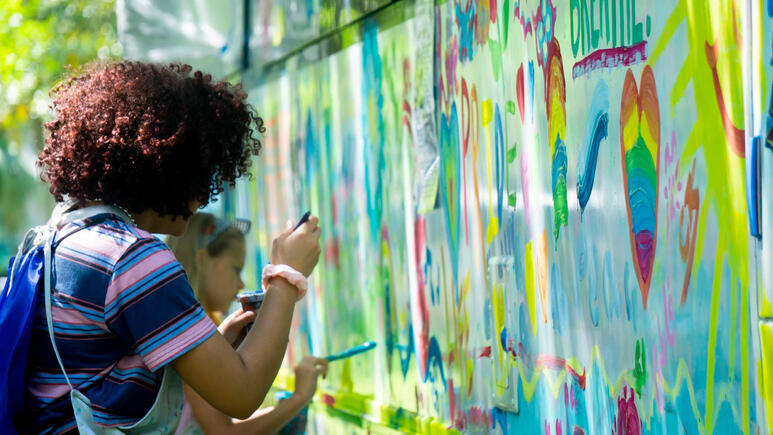 Students paint on a long wall, green splashes mixing in with red, in a summer day.