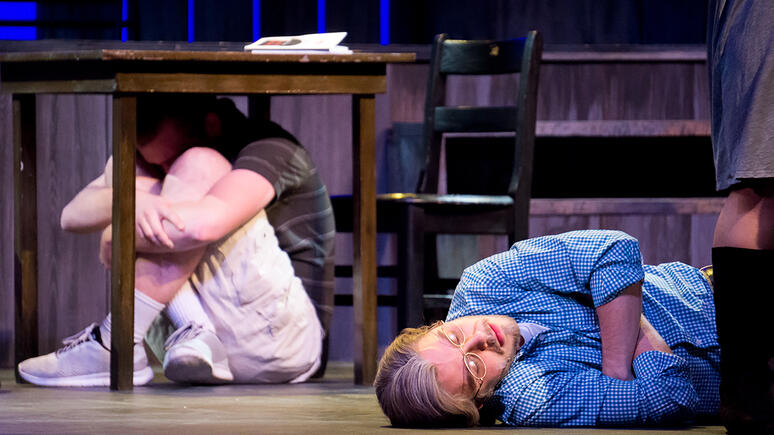 Two actors play dead on stage, one underneath a table in the fetal position, the other, arms crossed, sordid and exhausted expression on his face.