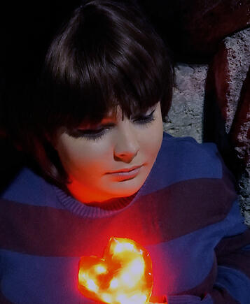A portrait shot of a cosplayer. It's dimly lit, warm and inviting, with a focus on a bright plastic heart glowing underneath a sweater.