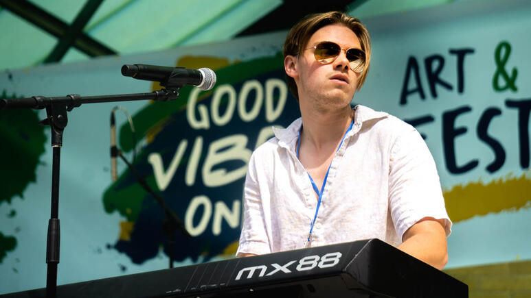 A keyboardist in a band plays under the bright sun, the event name a splash of color behind him.