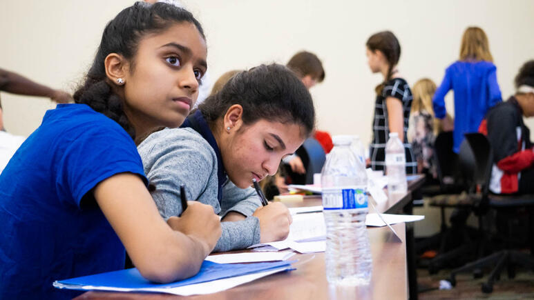 A row of students studying in a busy classroom. The focus is a young girl in the left, looking off toward the upper right, at an off-screen speaker. She's taking notes alongside her friend.