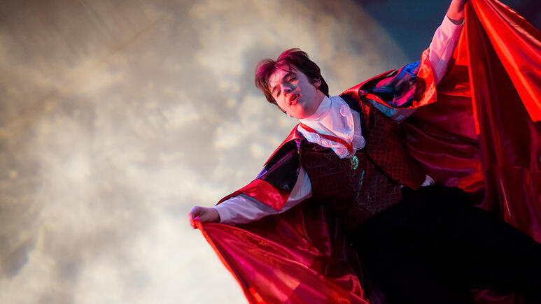 A performer, dressed in a Gothic Dracula-inspired outfit, raises his cape in a dramatic gesture against a full moon.