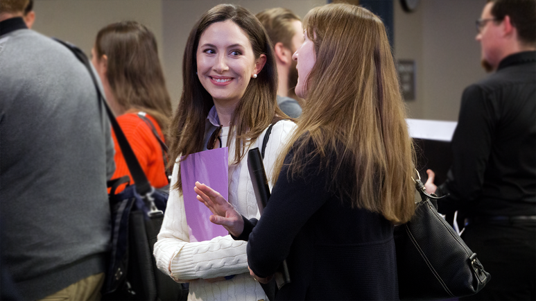Two students stand in the center, surrounded by a moving crowd, heading toward a presentation. They look at one another and talk vibrantly.
